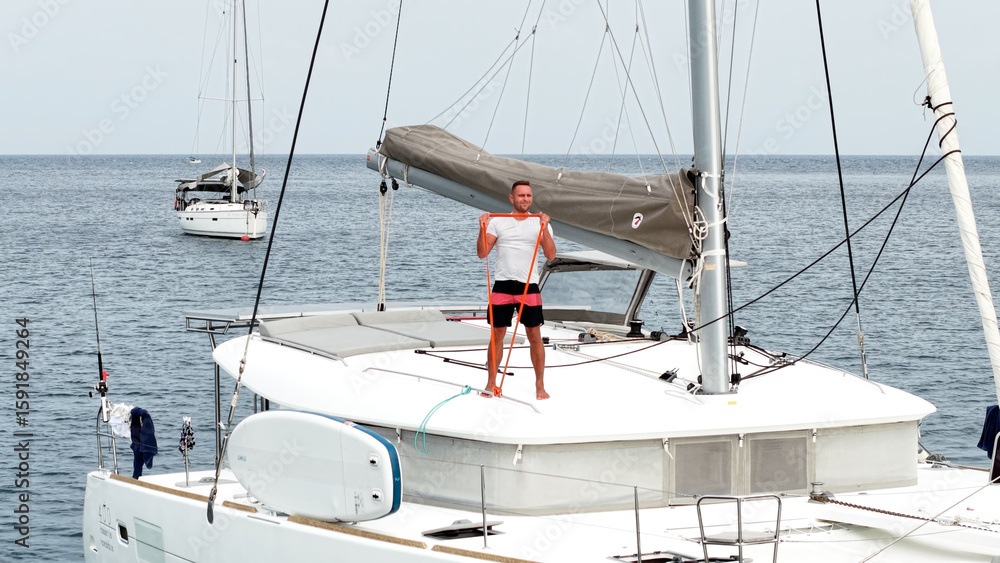 Morning on a yacht. Fitness exercises on the roof of a catamaran. A man doing exercises during a sea voyage. A man doing fitness exercises with a rubber elastic band Morning on a yacht. Fitness exercises on the roof of a catamaran. A man doing exercises during a sea voyage. A man doing fitness exercises with a rubber elastic band