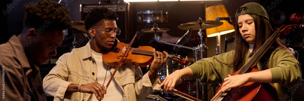 Fototapeta premium Black young adult man playing violin next to Latin young adult woman playing cello and Black young adult man playing keyboard in music studio, with drum set in background