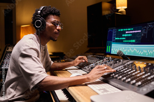 Fotografi Young adult Black man wearing headphones operating audio mixing console and comp