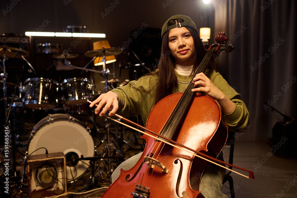 Fototapeta premium Portrait of Latin female musician sitting in music studio playing cello, holding bow and looking into camera, drum set and musical equipment visible in background