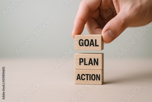 Hand stacking wooden blocks labeled goal plan action on a flat surface with a plain background.