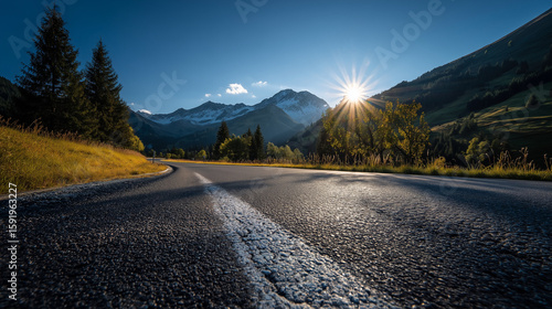 Foothills of the Austrian Alps with asphalt road on foreground and sun on blue morning sky
