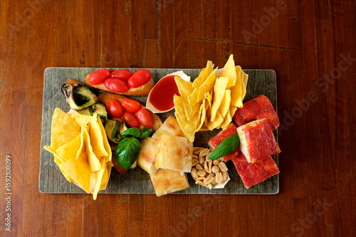 Tagliere misto per aperitivo con focaccia, bruschette, pomodorini, verdure grigliate, patatine, arachidi, salsa rossa e pizza al pomodoro. Vista dall’alto su tavolo in legno.