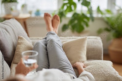 
Woman drinking water and resting with legs elevated on pillows, recovery self-care scene.
