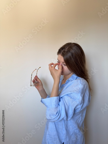 A young woman removes her glasses and pinches the bridge of her nose, a familiar gesture of eye strain from extended screen exposure or reading. digital eye strain, screen time fatigue