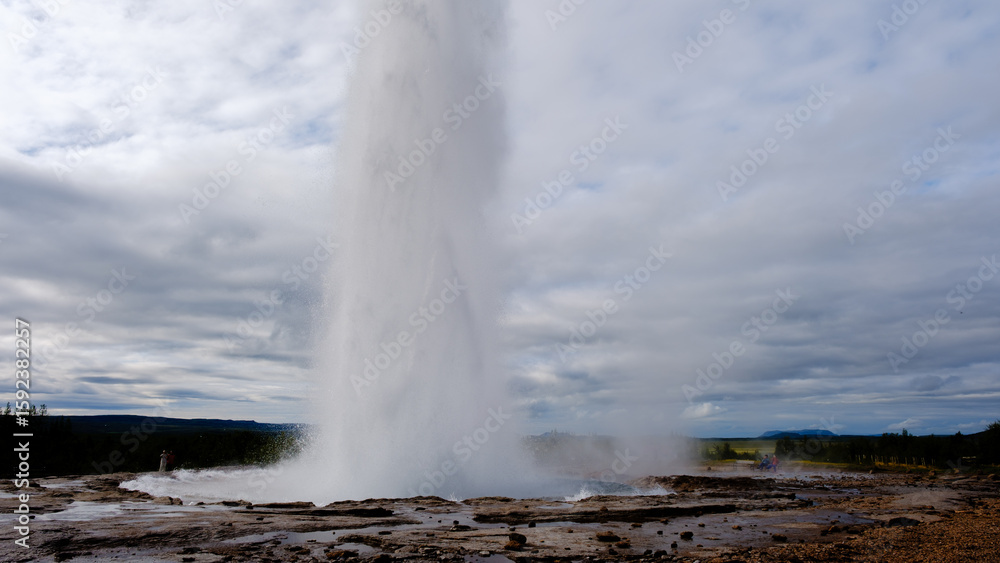 Obraz premium Strokkur geyser erupts powerfully into the air, surrounded by a rugged landscape in Iceland. Visitors marvel at this natural wonder, showcasing the beauty of geothermal activity in the region.