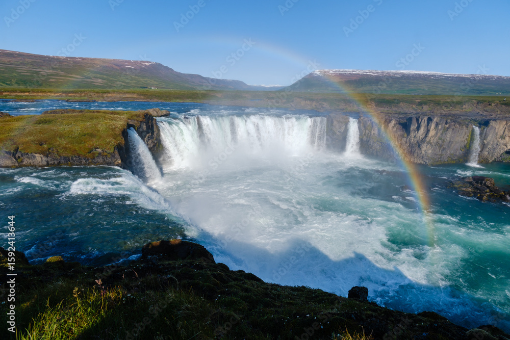Fototapeta premium Experience the stunning beauty of Godafoss waterfall plunging into turquoise waters, surrounded by lush greenery and a vibrant rainbow under a clear blue sky, showcasing natures essence in Iceland