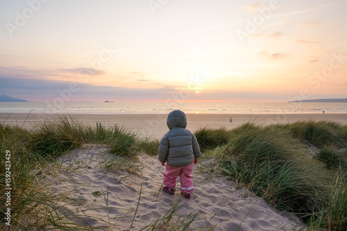 Child in a winter jacket standing on a sandy beach at sunset
