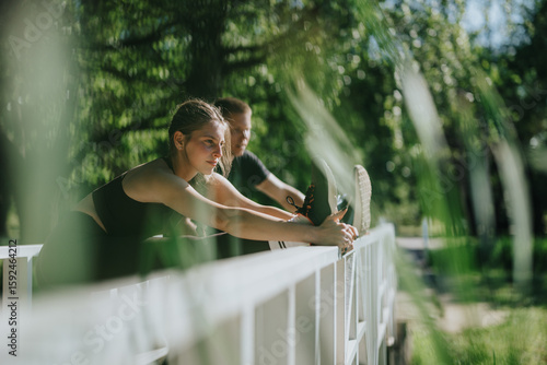 Couple practicing stretching exercises together on a bridge surrounded by lush greenery. The natural sunlight and serene atmosphere create a perfect setting for outdoor workouts and physically active.