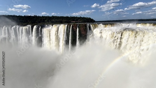 Panoramic view of Iguaçu magnificent and powerful waterfall with massive water flow, creating a dense mist and a rainbow below, surrounded by a lush green rainforest under a blue sky with clouds.