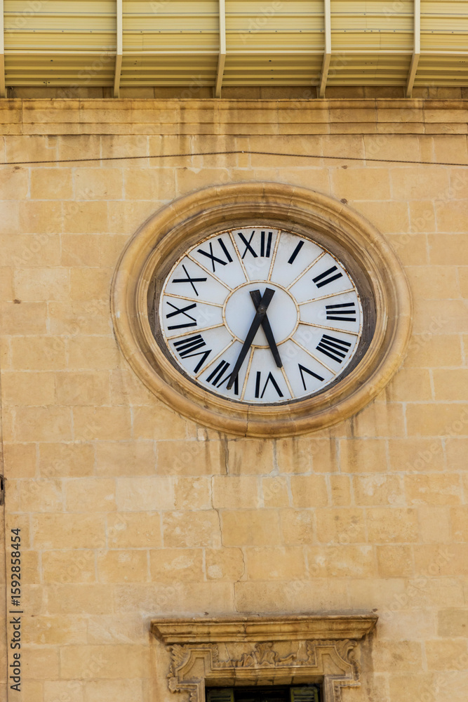 Fototapeta premium Close-up of a large clock with Roman numerals on a stone wall, showcasing intricate detailing and classic design. The clock is an architectural feature in Alicante, Spain.