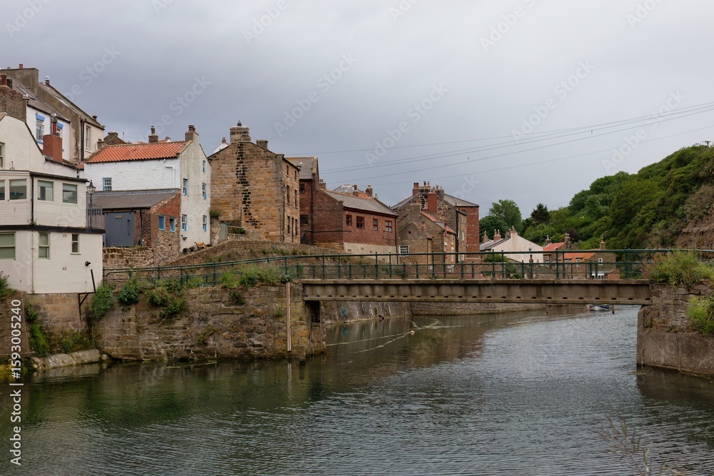 Fototapeta premium An outdoor shot showcases a body of water with buildings on the bank. The buildings are various colors and styles with a bridge over the water in Staithes - North Yorkshire - United Kingdom