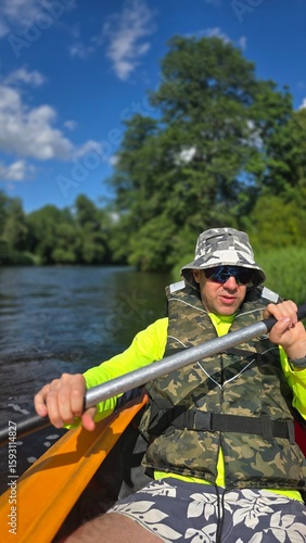 A man is kayaking, in the summer, holding an oar (wearing glasses, hat)