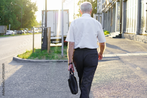 Elderly man walks along city street carrying a briefcase during late afternoon light in urban setting