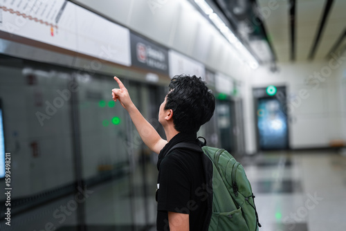 The image shows a young man, wearing a backpack, pointing at a map or information board, likely in a subway or metro station