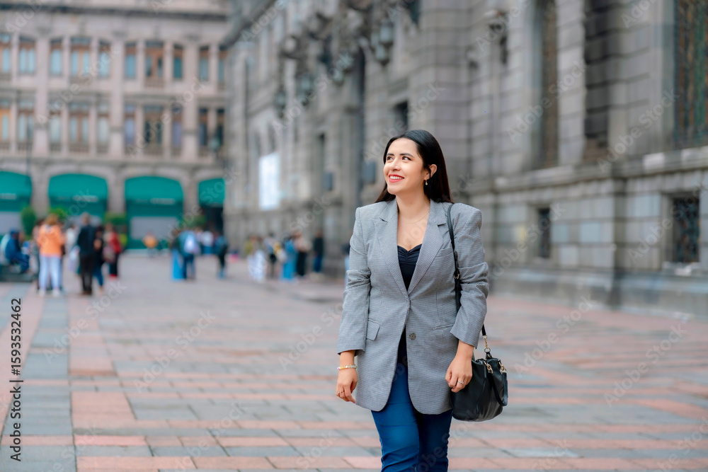 Fototapeta premium Confident young Latina business woman in gray blazer walking near MUNAL in Mexico City, smiling with historic architecture in the background