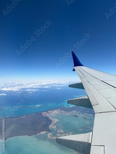 Stunning aerial view from airplane window showing tropical coastline with turquoise waters, small islands, and coral reefs beneath aircraft wing against clear blue sky during flight