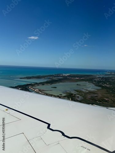 Breathtaking aerial view from airplane wing showing tropical island coastline with turquoise lagoons, coral formations, and pristine beaches under brilliant blue sky with scattered white clouds