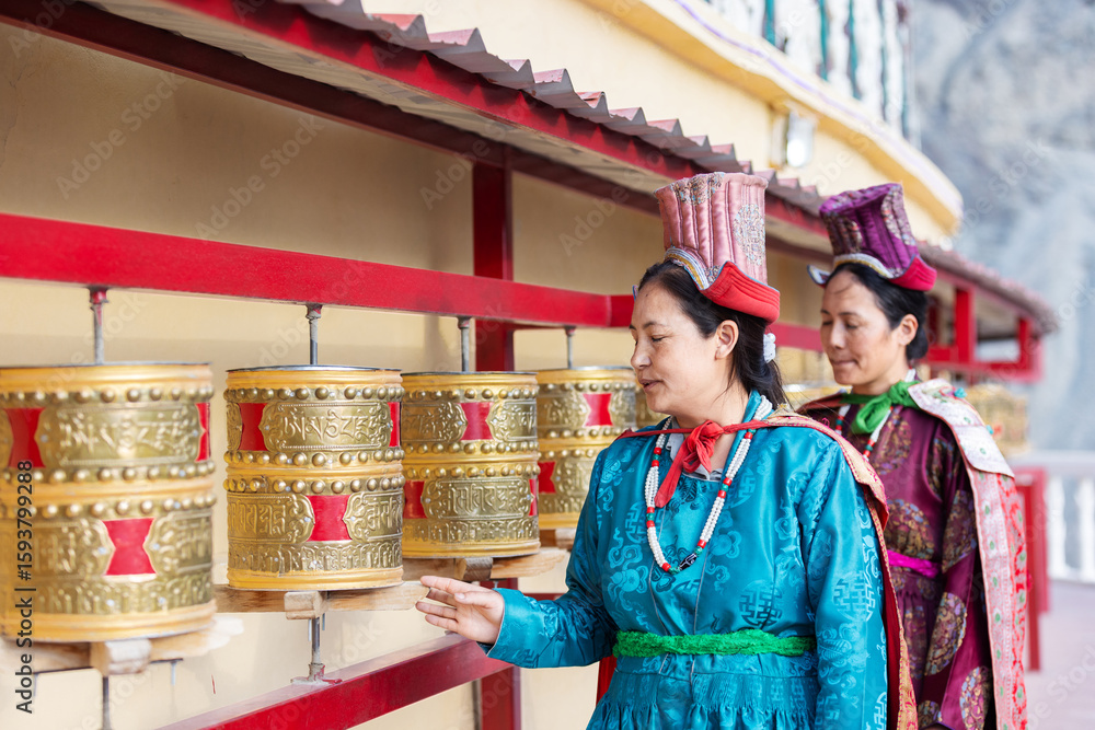 Fototapeta premium Two Indian women in traditional Leh attire spin golden prayer wheels at a monastery, expressing devotion and faith in a serene Himalayan setting, surrounded by rich colors and sacred symbolism.