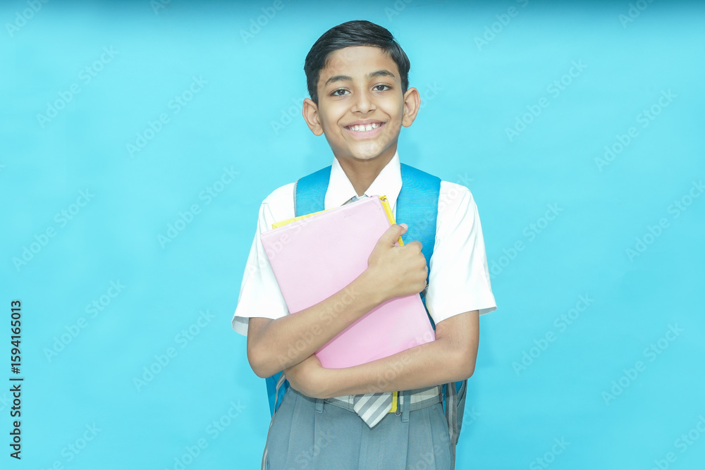 Obraz premium Happy Indian student schoolboy wearing uniform holding notebooks and bag standing isolated over blue background, Education knowledge concept. Copy space, Studio shot.