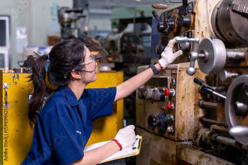 Closeup Asian woman industrial engineer in uniform checking old model lathe and drilling machine with clipboard at industrial workshop.