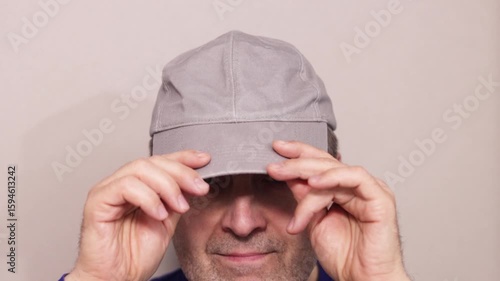 A cheerful, elderly, unshaven man puts on a cap with both hands, looks at the camera and makes a positive gesture with a thumb.