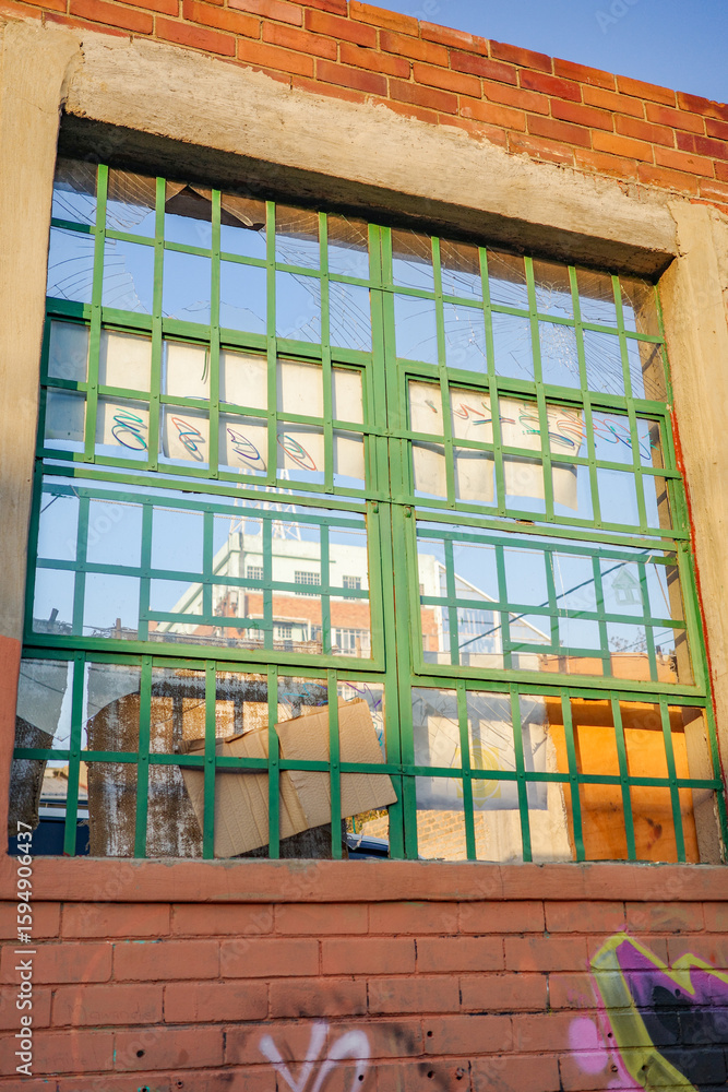 Obraz premium Close-up shot of a green metal-barred window on a worn-out urban brick building, featuring broken glass, graffiti, and cardboard covering the interior. Abandoned industrial aesthetic