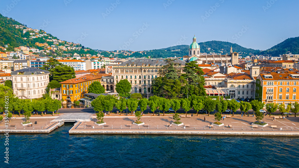 Naklejka premium Aerial view of Como village with clustered terracotta rooftops, narrow streets, lakeside promenade, and surrounding green hills under clear skies