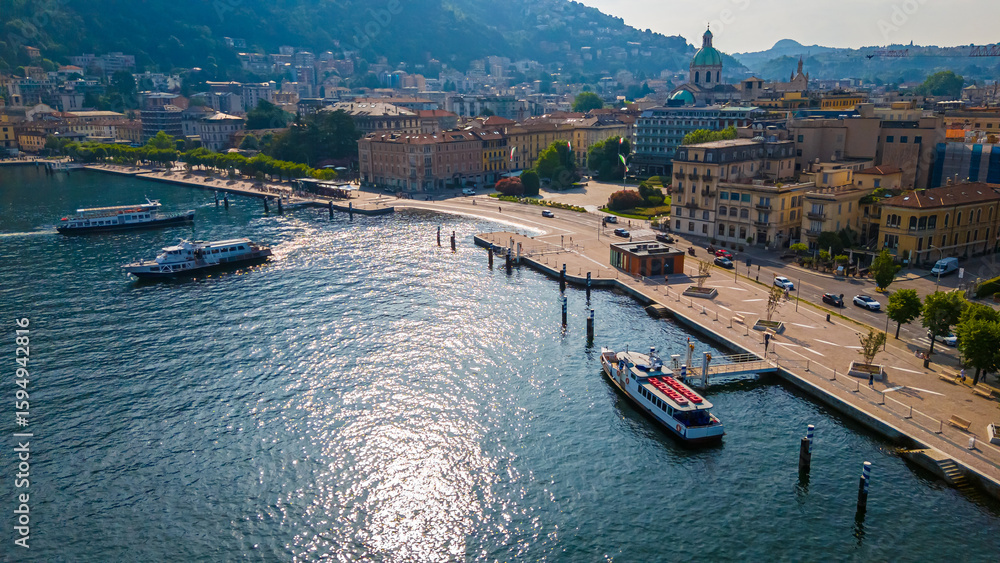 Fototapeta premium Aerial view of Como village with clustered terracotta rooftops, narrow streets, lakeside promenade, and surrounding green hills under clear skies