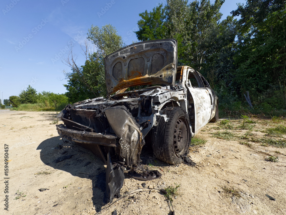 Fototapeta premium A heavily burned-out car with its hood open sits abandoned on a dirt path under a clear sky, surrounded by green trees and bushes.