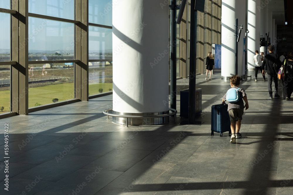 Fototapeta premium A child walks away from the camera in a sunlit airport terminal, pulling a small suitcase. The interior features tall windows casting long shadows on the polished floor.