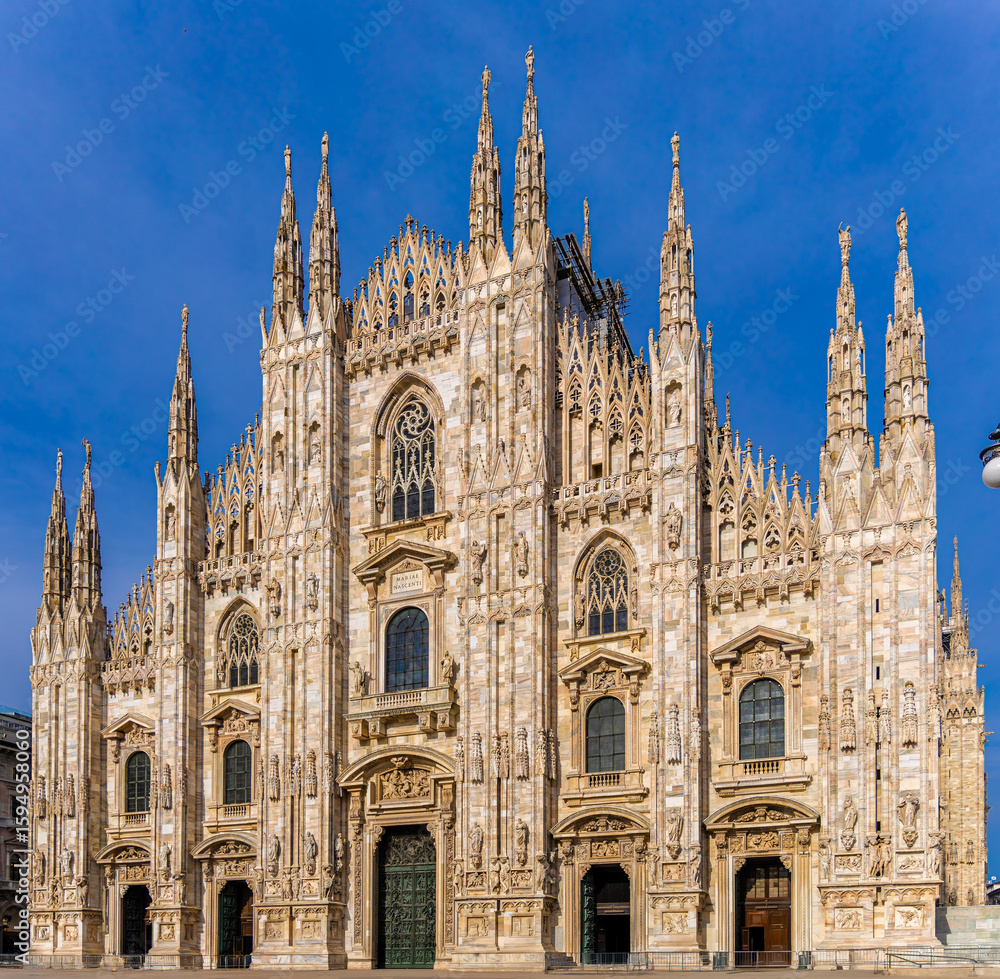 Gothic-style Milan Cathedral facade with ornate spires and intricate stonework against clear blue sky, captured in daylight Gothic-style Milan Cathedral facade with ornate spires and intricate stonework against clear blue sky, captured in daylight