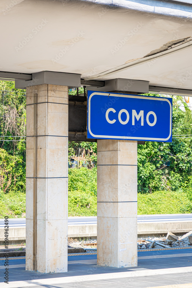 Fototapeta premium Como train station platform in Italy with overhead sign reading “Como,” railway tracks, and shelter under a bright summer sky