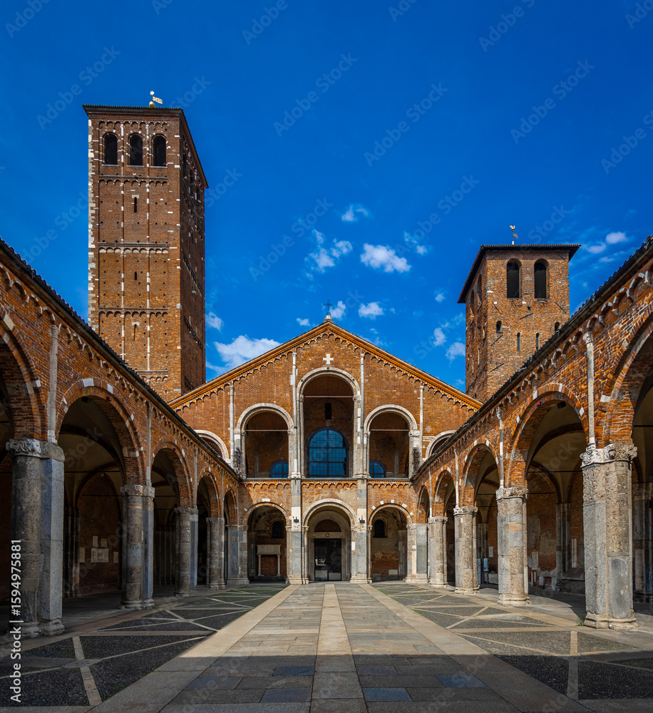 Obraz premium Courtyard view of Basilica di Sant’Ambrogio in Milan with Romanesque arches, brick facade, and twin bell towers under a vivid blue sky