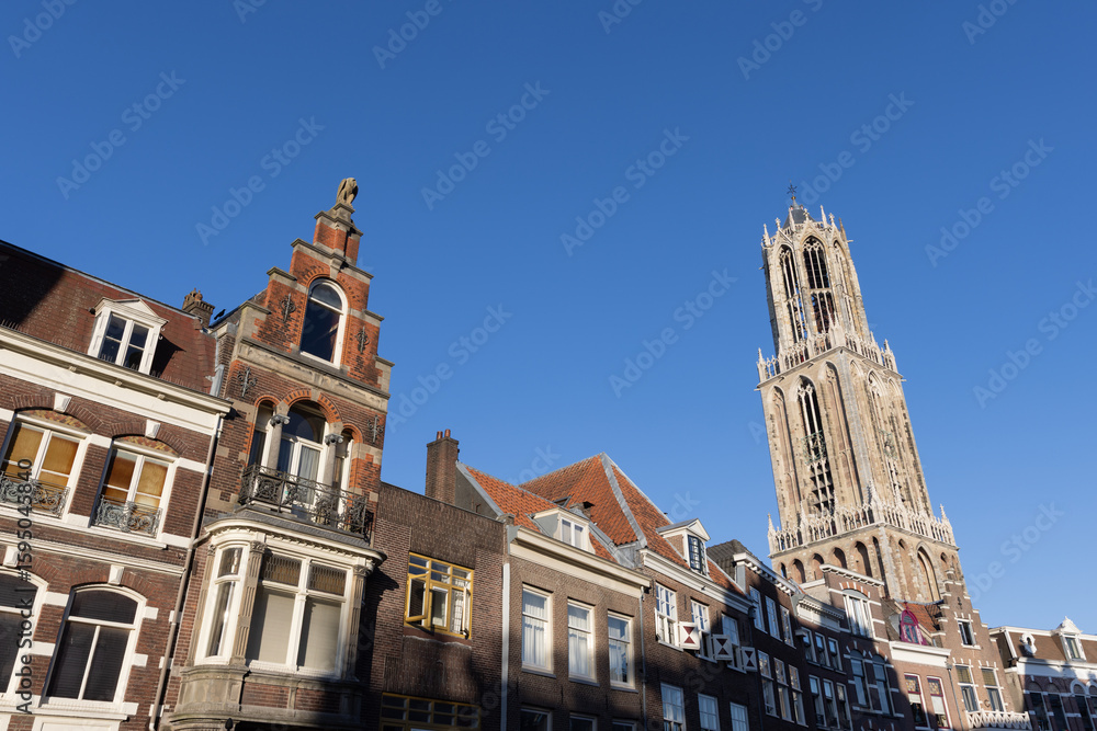 Obraz premium A classic low-angle view showcasing the varied gabled rooftops of historic Dutch houses leading up to the impressive Dom Tower of Utrecht Dom toren against a brilliant blue sky.