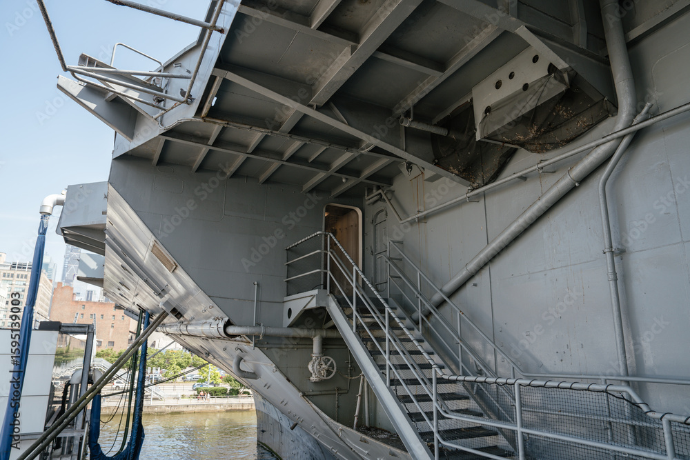 A detailed view of a metal staircase and exposed piping along the gray hull of a docked aircraft carrier. The structure shows industrial design elements, railings, and mooring lines. A detailed view of a metal staircase and exposed piping along the gray hull of a docked aircraft carrier. The structure shows industrial design elements, railings, and mooring lines.
