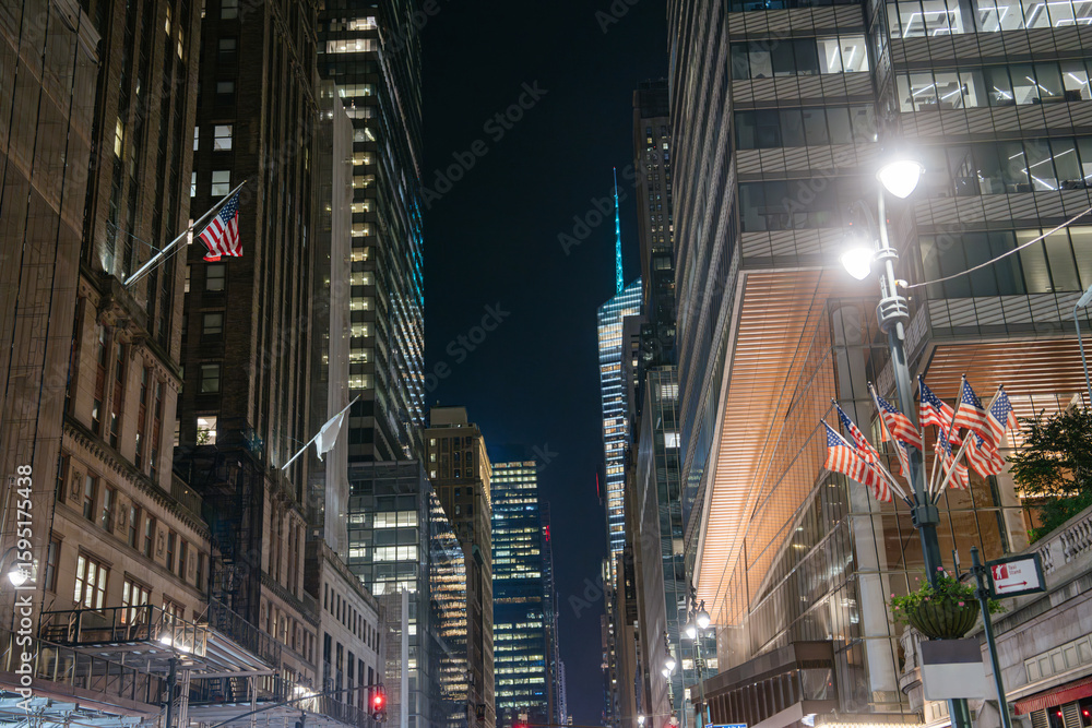 Obraz premium Multiple American flags line the street between tall Midtown skyscrapers glowing under the night sky. The scene is lit by streetlamps and reflects the patriotic spirit of New York City.