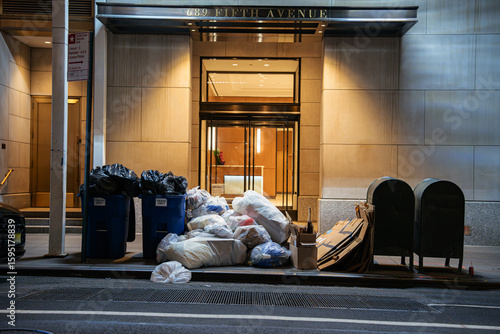 A large pile of garbage bags and flattened cardboard boxes sits near the entrance of a building on Fifth Avenue in New York City. The scene contrasts luxury architecture with urban waste visible on