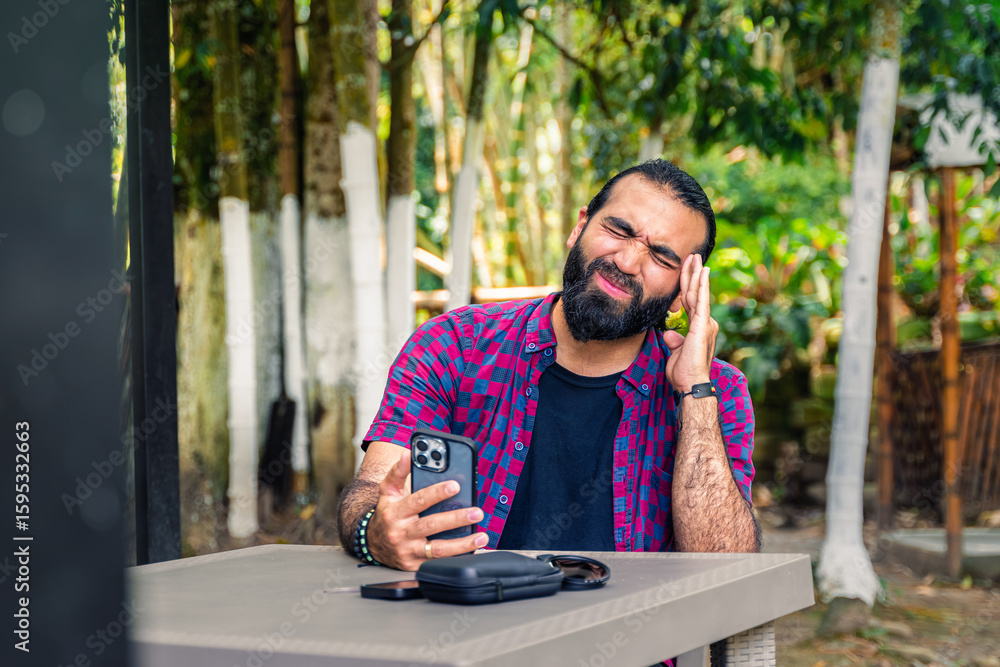 Fototapeta premium Stressed Colombian man in plaid shirt holding phone and pressing hand to forehead during remote work in tropical garden in Santuario, Risaralda, with space for text