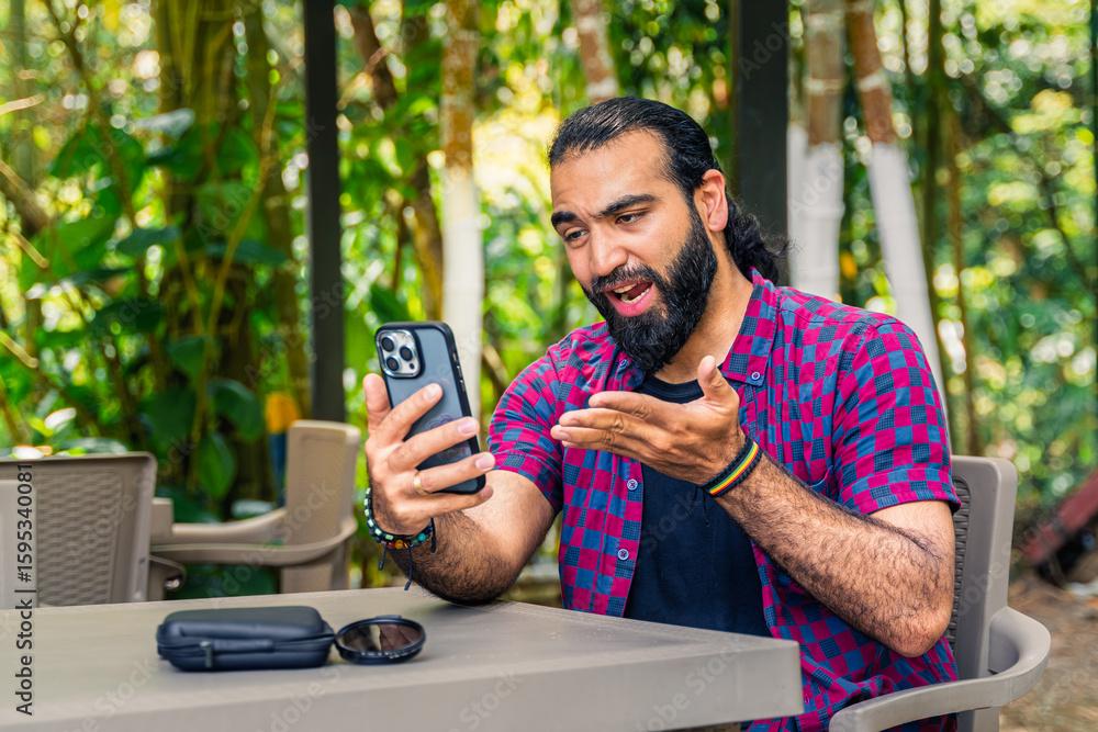 Expressive Colombian man in plaid shirt reacting with dissatisfaction while holding phone at outdoor table in lush tropical garden in Santuario, Risaralda, with space for text. Expressive Colombian man in plaid shirt reacting with dissatisfaction while holding phone at outdoor table in lush tropical garden in Santuario, Risaralda, with space for text.