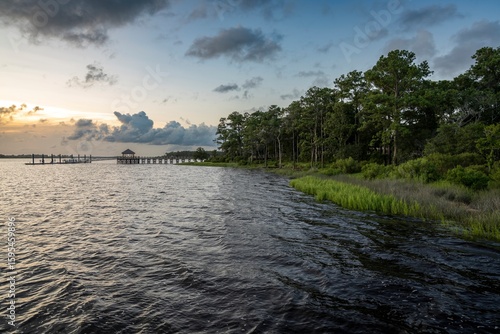 A peaceful North Carolina waterfront at sunset features a wooden pier extending into the water, bordered by lush green marsh grasses and tall pine trees under a partly cloudy sky.