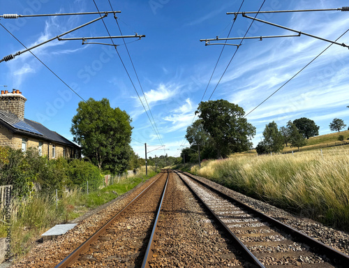 Ilkley to Leeds and Bradford railway tracks stretch into the distance under a clear blue sky, flanked by lush green trees and a quaint stone house on the left in, Burley in Wharfedale, Yorkshire, UK