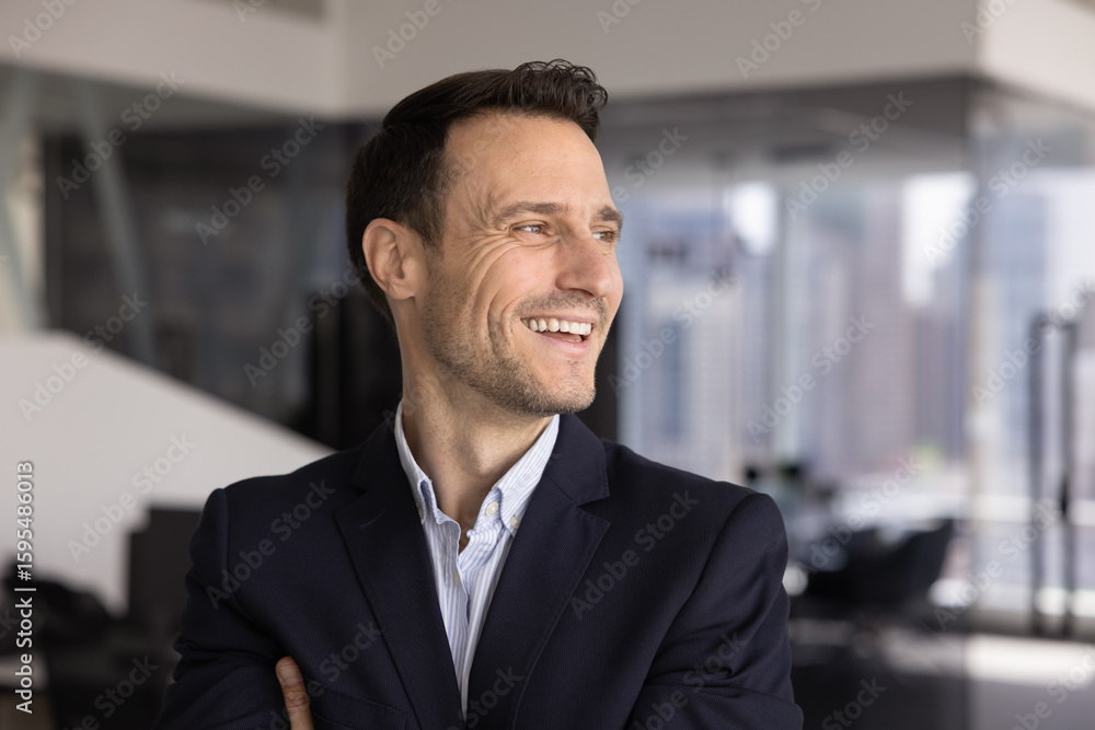 Fototapeta premium Head shot of middle-aged businessman posing in modern workplace, looks into distance with joyful and contented expression, enjoys career growth, feel proud and satisfied with professional achievements