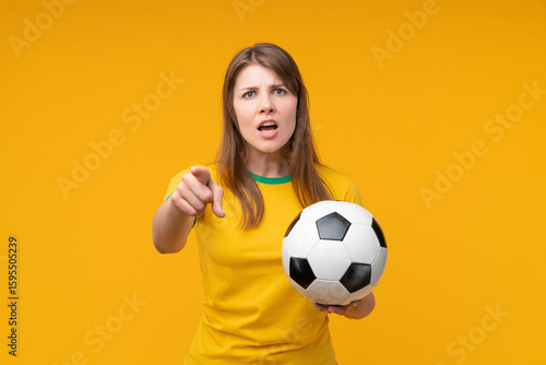 Confident young female soccer fan or player holding a soccer ball and pointing at the camera with an accusing expression on yellow background