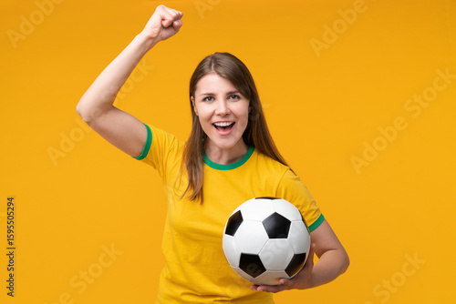 Happy young woman in yellow shirt cheering with raised fist and holding soccer ball
