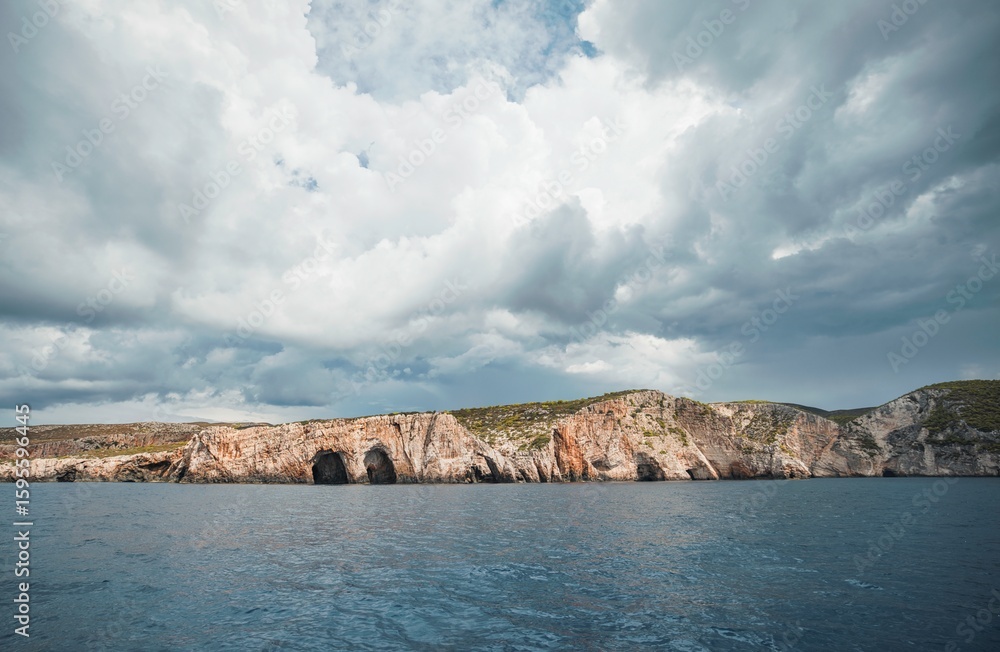 Naklejka premium A boat tour moves along the Ionian Sea near Zakynthos. Caves are visible in the rocky coastline as the sky churns above, promising rain