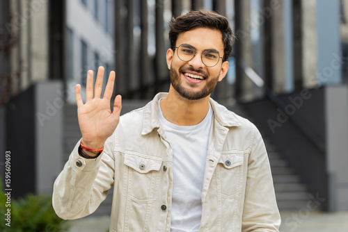 Hello. Indian man smiling friendly at camera, waving hands gesturing hi, greeting or goodbye welcoming with hospitable expression outdoors. Excited Arabian Hindu guy standing in urban city town street