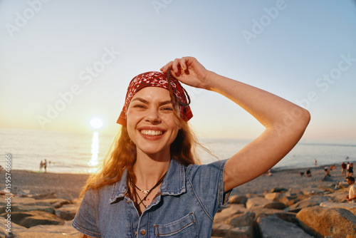 Vászonkép smiling woman wearing denim jacket and red bandana on beach during sunset, boho-
