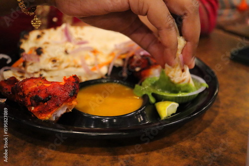 Indian street food close-up – Hand dipping naan into green chutney with tandoori chicken, dal, and lime on traditional platter – Authentic South Indian meal, vibrant and flavorful.