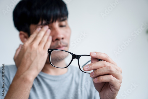 Tired Asian Man Experiencing Eye Pain and Blurry Vision, Holding His Eyeglasses in Hand While Struggling with Eye Strain and Fatigue